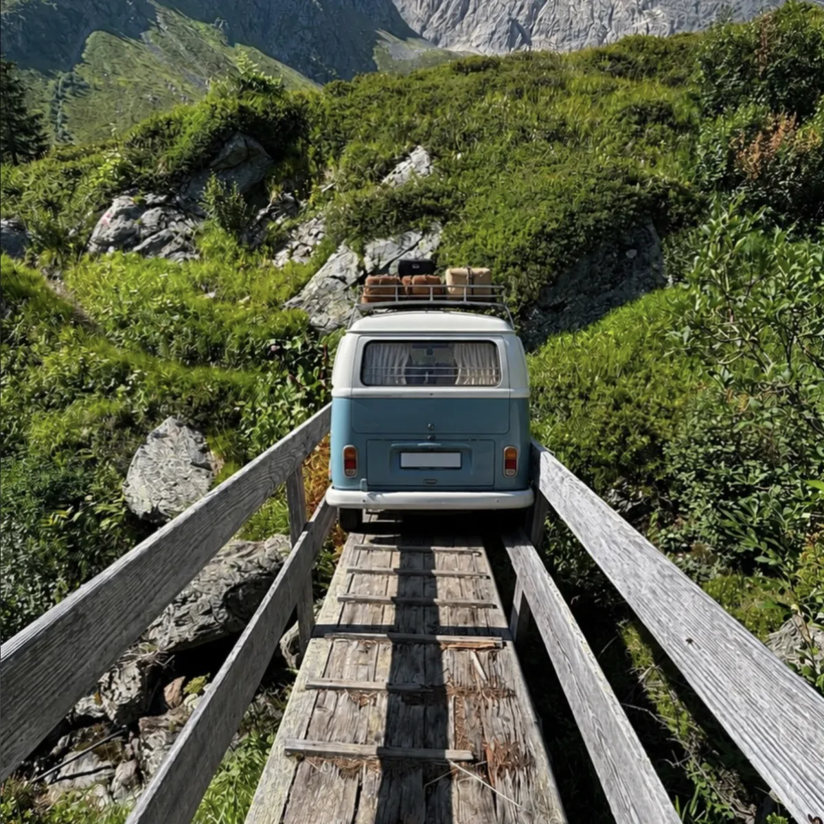 Ein alter Bulli fährt über eine schmale Holzbrücke in einer sommerlichen Berglandschaft